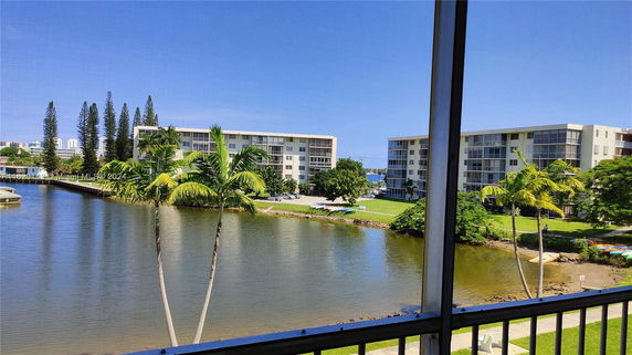 View of two mid-rise apartment buildings near a waterfront area with palm trees and greenery.