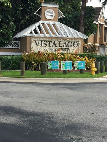 Front view of a building entrance labeled 'Vista Lago' with a decorative roof and surrounding greenery.