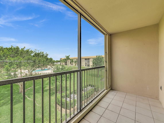 Balcony view overlooking a garden area with trees and a distant swimming pool.
