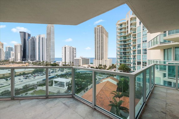 Panoramic view from a balcony of a high-rise building overlooking a city skyline with other tall buildings and the ocean in the distance.