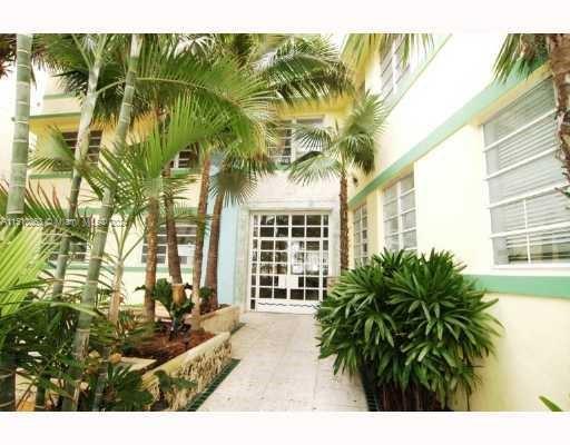 Front view of a multi-story building with light yellow exterior and white framed windows surrounded by tropical plants.