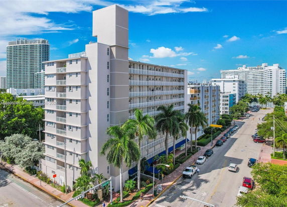 Front view of a multi-story apartment building with balconies and palm trees lining the street.