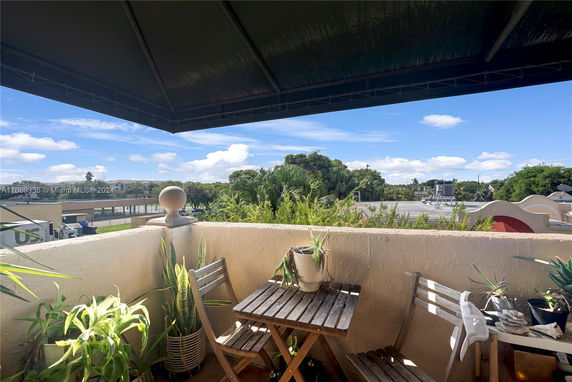View from a balcony with potted plants and wooden furniture, overlooking trees and a flat rooftop.