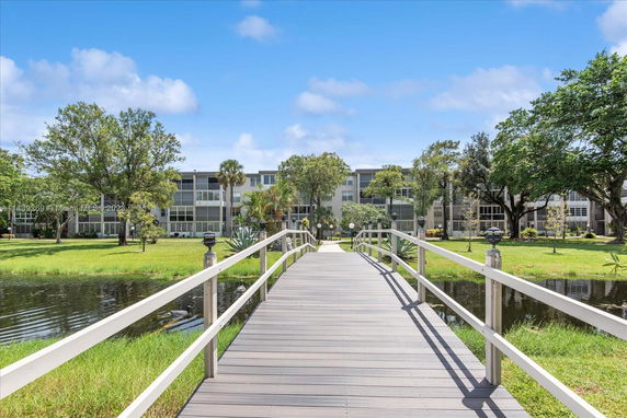 Exterior view of a multi-story residential building with a wooden bridge over a small body of water and surrounding greenery.
