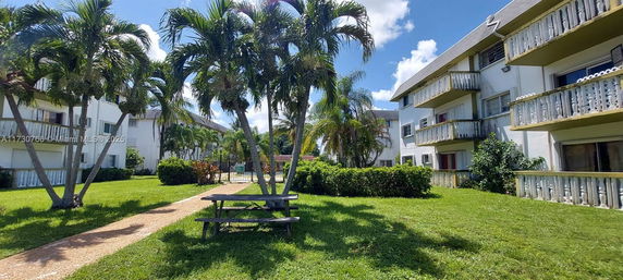 Outdoor view of residential buildings with balconies and a pool area surrounded by palm trees.