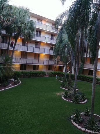 View of a multi-story residential building with balconies, surrounded by palm trees and a neatly landscaped courtyard.
