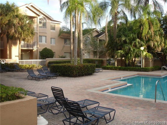 Outdoor swimming pool area with surrounding lounge chairs and a multi-story apartment building in the background.