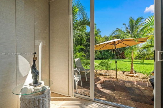View through a glass door of a patio with chairs and an umbrella, surrounded by lush greenery.