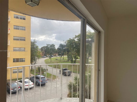 View from a balcony showing a nearby yellow building, parked cars, and a grassy area with trees.