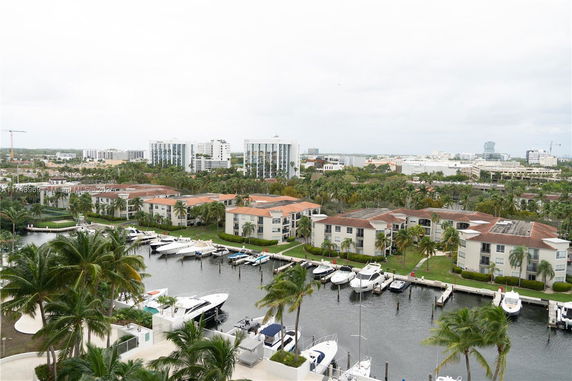 Aerial view of buildings and boats docked at a marina, surrounded by palm trees and urban structures.