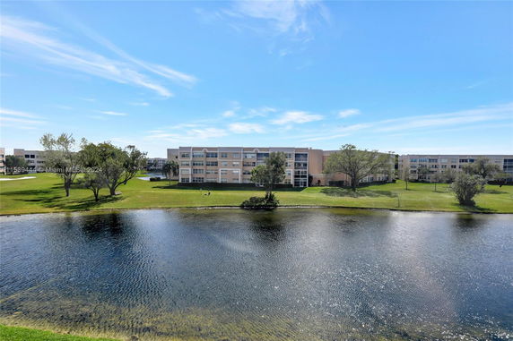 Wide view of residential buildings seen across a pond with a grassy area and trees in the foreground.