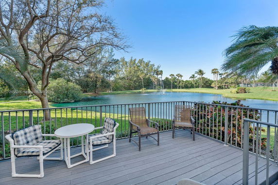 Outdoor seating area on a deck overlooking a pond with fountains and trees in the background.