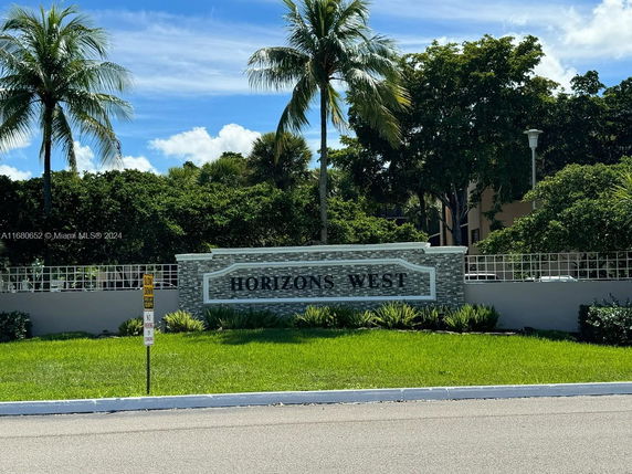 Front view of a property entrance sign with palm trees and surrounding greenery.