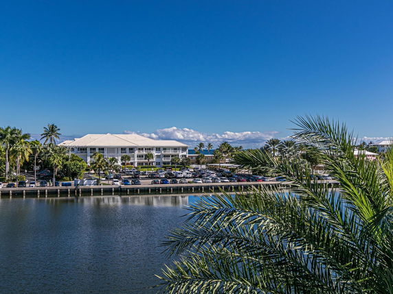 View of a large white building near a body of water with parked cars and palm trees in the vicinity.