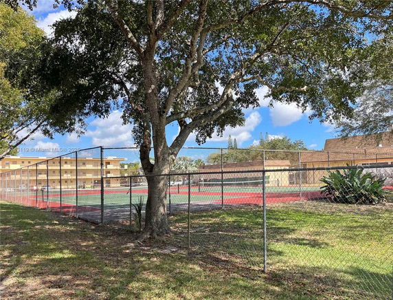 View of a fenced tennis court with surrounding trees and residential buildings nearby.