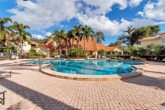 Outdoor swimming pool area with a large pool and surrounding patio near a residential building with a red-tiled roof.
