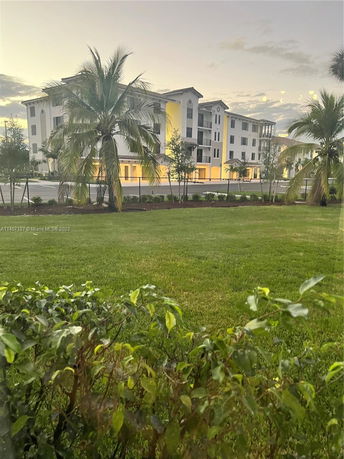 Distant view of a multi-story residential building with balconies and large windows, surrounded by palm trees and greenery.
