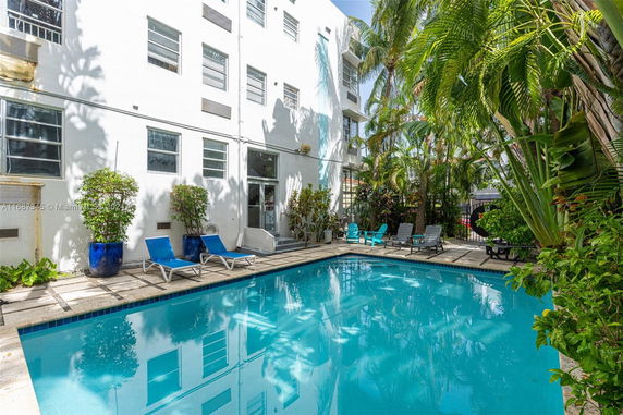 Outdoor swimming pool area adjacent to a multi-story white building with lounge chairs and tropical plants.