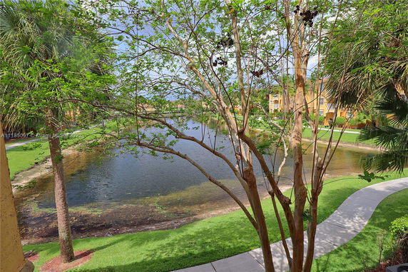 View of a lake surrounded by green grass and trees, with buildings visible in the background.
