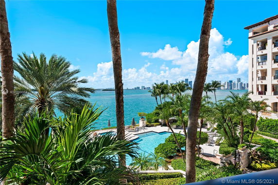 View of a waterfront area with palm trees and a pool, adjacent to a multi-story building.