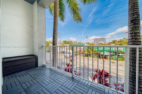 View from a balcony overlooking a street and nearby buildings with palm trees and clear skies.