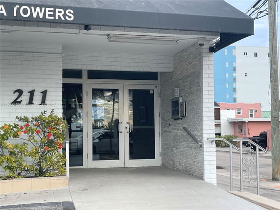 Entrance of a building with white brick walls and glass doors, showing the number 211 and a black awning above.