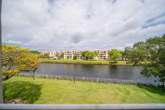 View of residential buildings across a water body with trees and grassy area in the foreground.