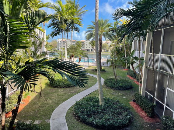 View of a courtyard area with palm trees between two apartment buildings, with a swimming pool visible in the background.