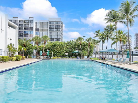 Swimming pool area with a high-rise building in the background, surrounded by palm trees and lounge chairs.