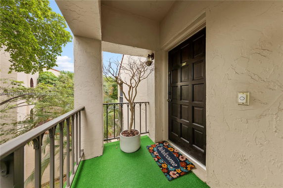 View from a balcony with a welcome mat and a potted plant, overlooking trees and exterior of nearby buildings.