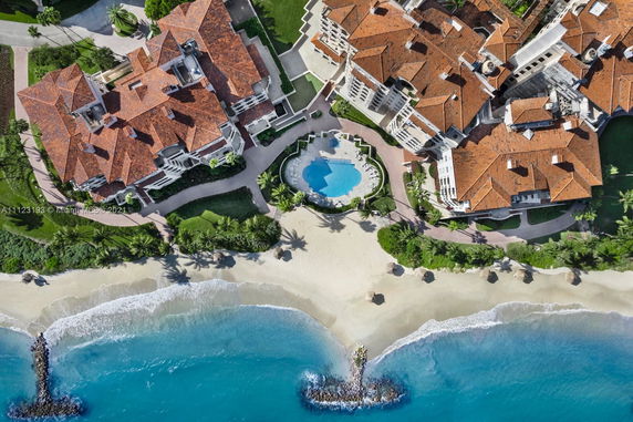 Aerial view of buildings with red-tiled roofs adjacent to a sandy beach and pool area.
