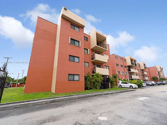 Front view of a multi-story apartment building with balconies and a parking area.