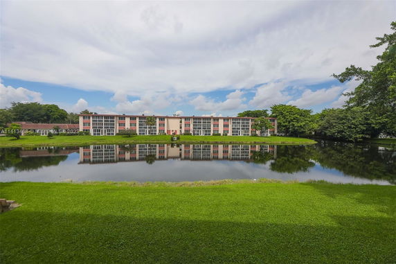 A three-story building overlooking a pond with green grass and trees around.