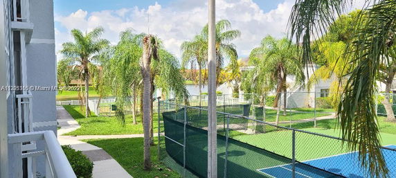 View of an outdoor tennis court surrounded by palm trees and nearby buildings.