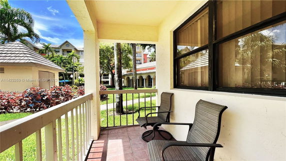 View from a covered patio area with two chairs overlooking a courtyard with gardens and a building in the background.