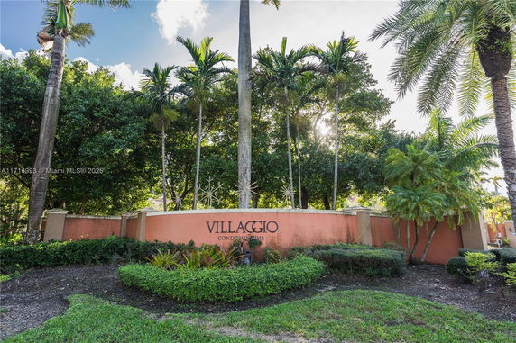 Front view of a condominium entrance with a sign reading 'VILLAGGIO Condominiums' surrounded by palm trees and bushes.
