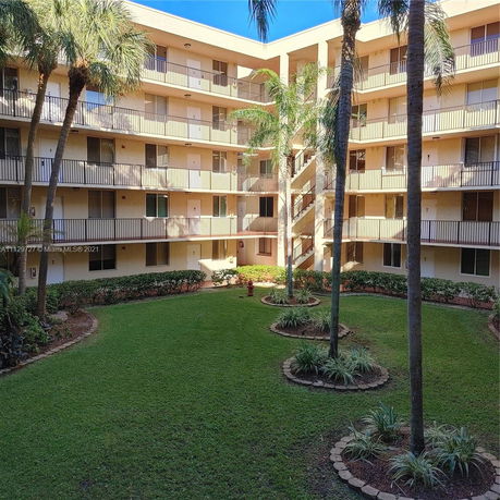 View of a multi-story apartment building surrounding a courtyard with grass and palm trees.
