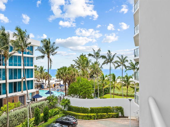 View from building balcony overlooking a pool area, palm trees, and the ocean in the background.
