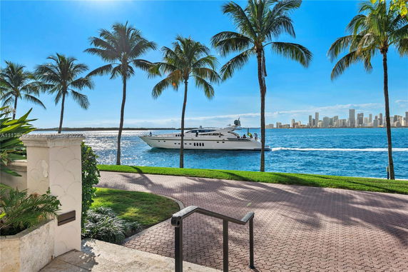 Scenic view of a waterfront area with a yacht passing by, palm trees, and a city skyline in the distance.