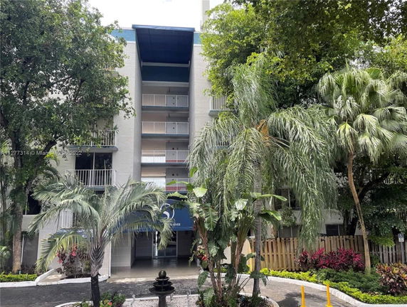 Front view of a multi-story residential building with balconies, surrounded by trees and greenery.