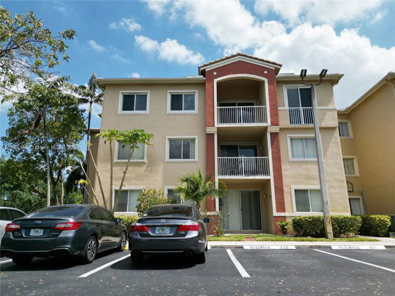 Front view of a three-story apartment building with balconies and two parked cars.