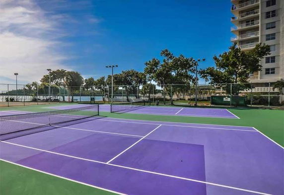 Tennis courts adjacent to a tall building with trees and a blue sky in the background.