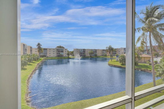 View of buildings across a lake with a fountain, surrounded by palm trees and grass.