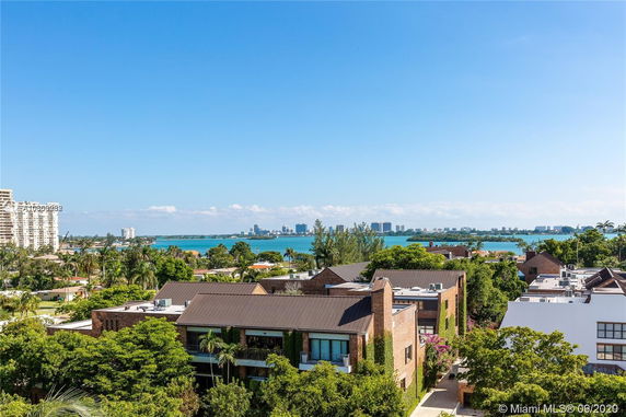 Aerial view of modern apartment buildings with brick exteriors and flat roofs, set amidst greenery, overlooking a body of water with a city skyline in the distance.