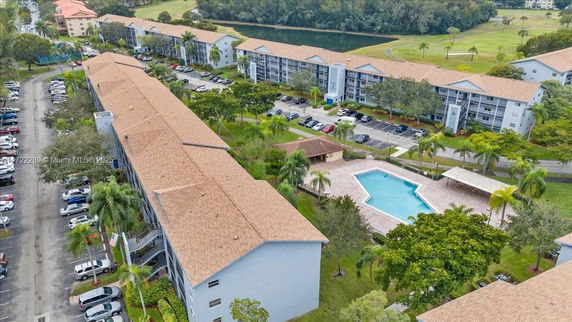 Aerial view of apartment buildings with a swimming pool, parking area, and greenery surrounding the complex.