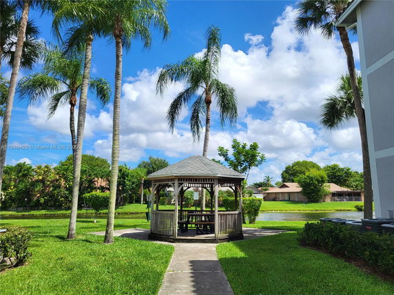 A gazebo in a well-maintained garden area with palm trees and a small pond nearby, adjacent to residential buildings.