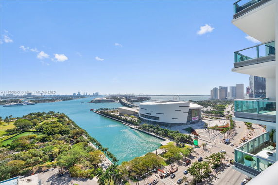 Aerial view of a waterfront area with buildings, trees, and a large body of water under a clear blue sky.