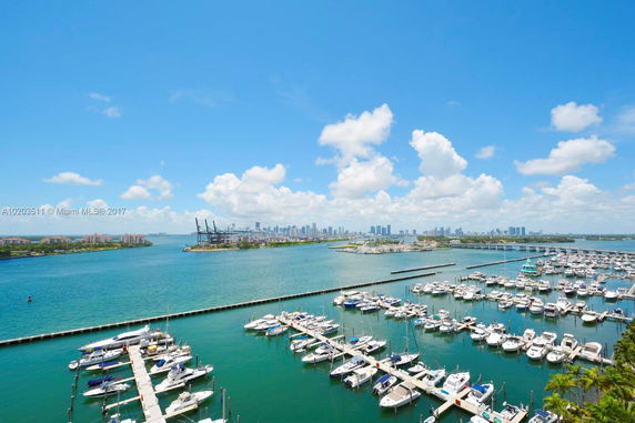Wide-angle view of a marina with numerous boats docked, and a distant city skyline across the water under a blue sky with scattered clouds.