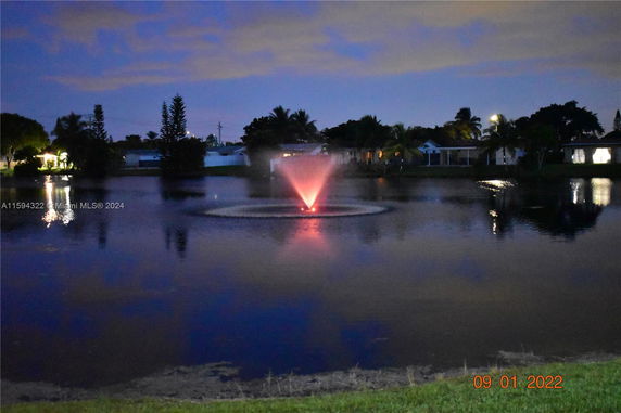 Nighttime view of a lake with illuminated fountain and houses in the background.