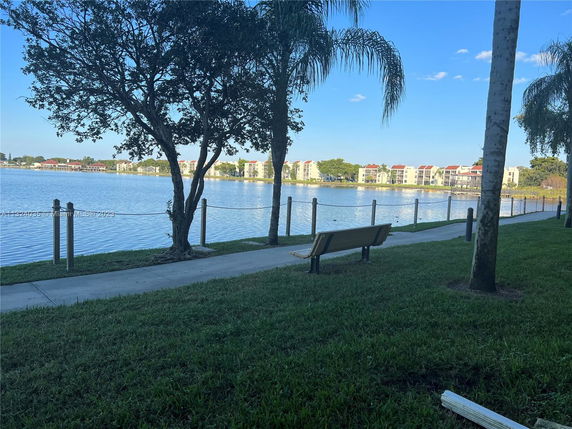 View of residential buildings across a lake with a pathway and bench in the foreground.